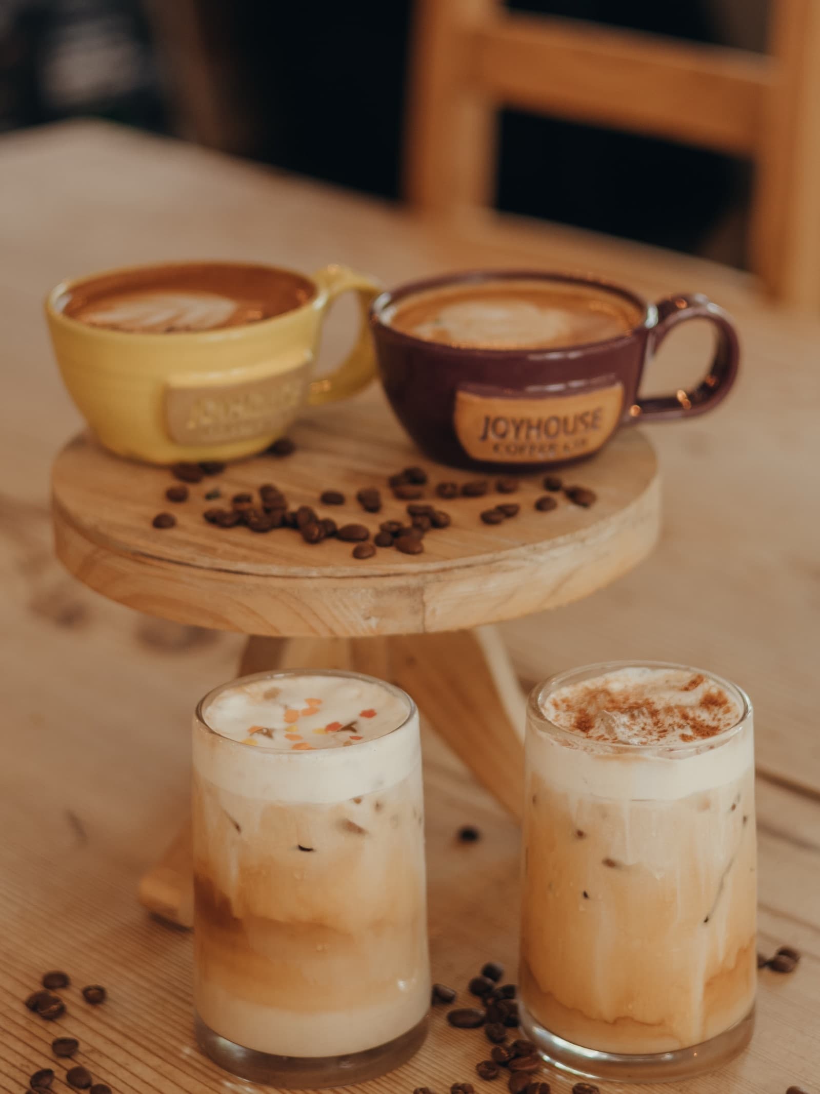Two lattes in Joyhouse mugs on a wooden stand scattered with beans, iced lattes below